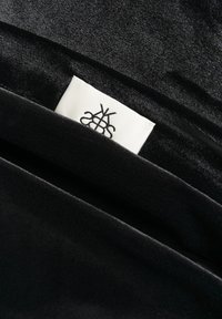 Must mustri materjal, millel on väikesel valgel sildil osaliselt nähtav must abstraktne logo.