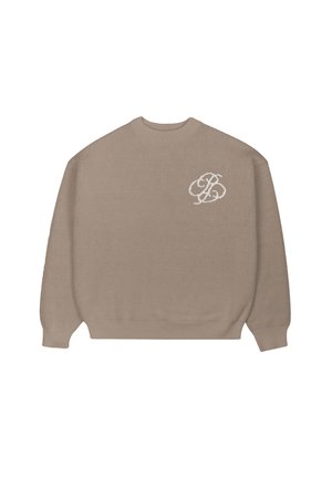Kobieta ubrana w brązowy sweter z białym logo, szare spodnie, pod spodem koszula w paski i brązowo-biała czapka podszyta polarem.