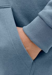 Material svetlo modrega puloverja z rebrastim manšetom, ki vključuje stranski žep. Gladek tekstura z vidnimi šivi in detajli šivanja.