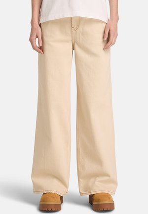 Beige bukser med brede ben lavet af robust bomuld med synlige syningsdetaljer, kombineret med brune plateau-sko. Bukserne har et design med høj talje.