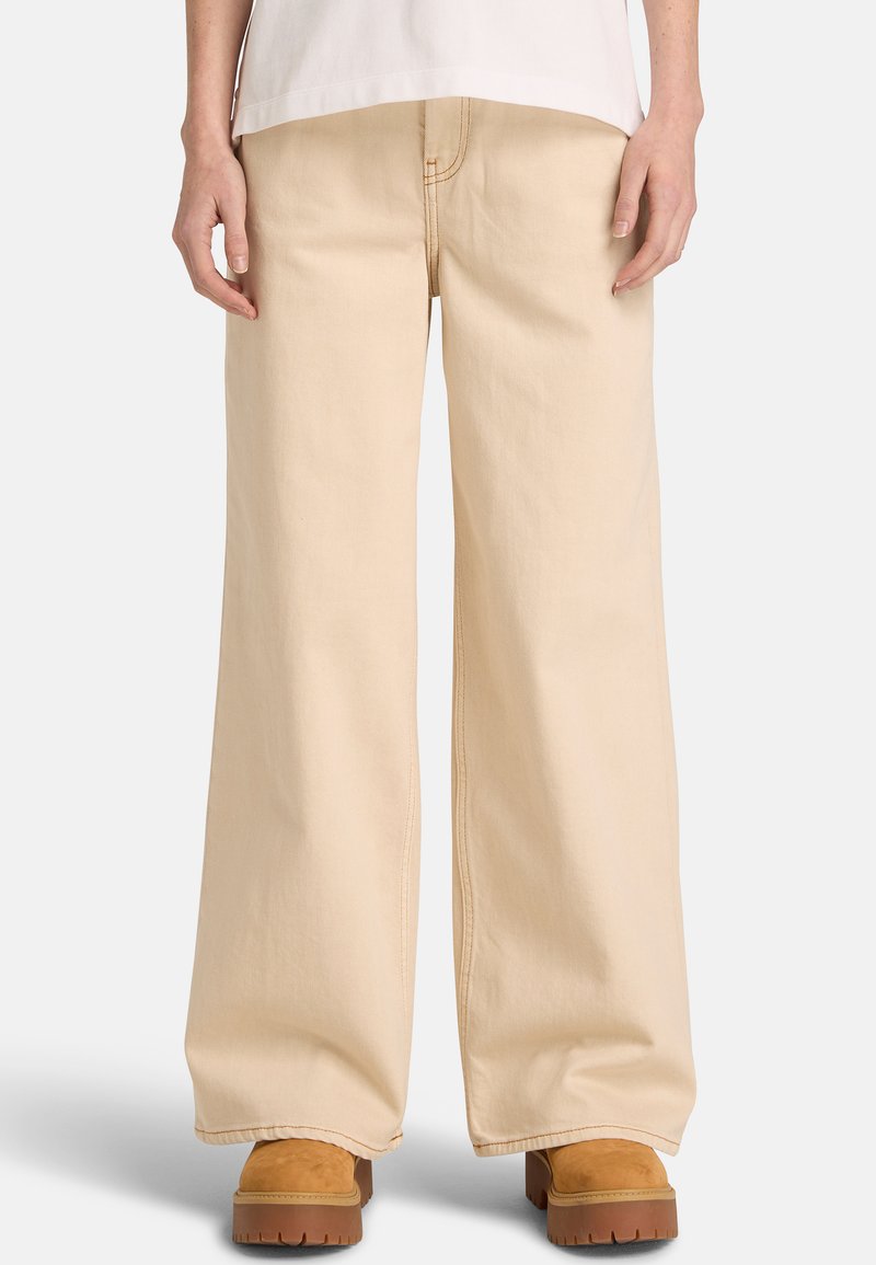 Pantalon large beige en coton robuste avec des détails de couture visibles, associé à des chaussures à plateforme marron. Le pantalon présente une coupe taille haute.