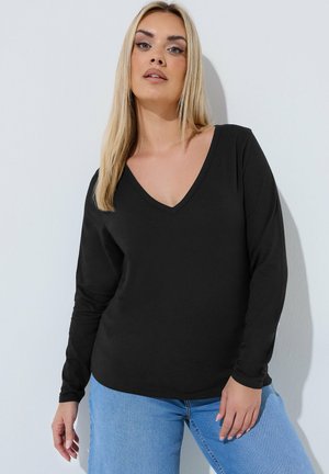 Kobieta o długich blond włosach, ubrana w czarną bluzkę z długim rękawem i dekoltem w serek oraz jasnoniebieskie dżinsy, stojąca przy białej ścianie.