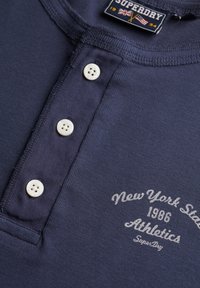 Close-up van een donkerblauw Henley-shirt met drie witte knopen en wit bedrukt tekst "New York State 1986 Athletics Superdry."