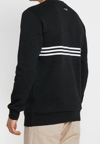 Svart sweatshirt i mjukt tyg med rund halsringning och tre horisontella vita ränder över ryggen. Normal passform, ribbad mudd.