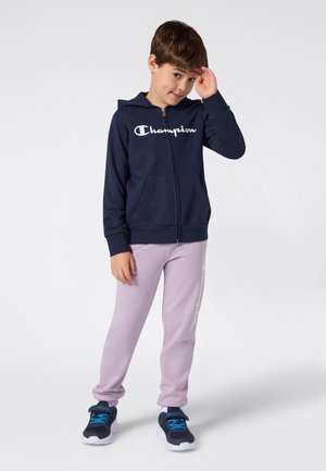 Marineblauer Kapuzenpullover mit durchgehendem Reißverschluss und weißem Logo, kombiniert mit hellvioletten Jogginghosen mit seitlichem Aufdruck. Kind trägt schwarze Sneakers mit blauen Akzenten.