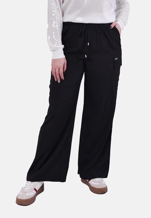 Persona che indossa pantaloni neri a gamba larga con coulisse, sneakers bianche con suola marrone e maglia bianca a maniche lunghe con dettagli in paillettes.