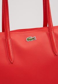Sac à main rouge texturé avec deux anses d'épaule et un petit logo crocodile vert près du centre supérieur.