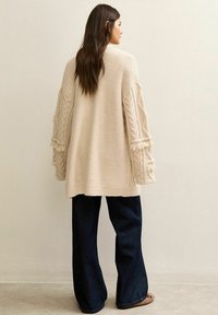 Crème kabelgebreide cardigan met franjes en brede mouwen, gecombineerd met donkerblauwe wijde jeans. Gladde textuur, ontspannen pasvorm.