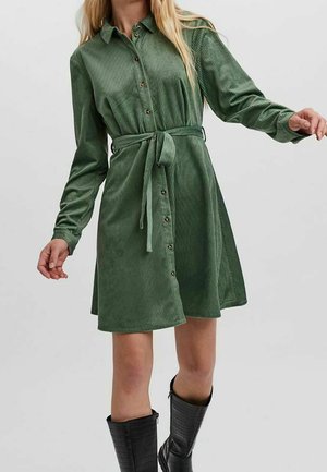 Woman wearing a knee-length, green corduroy shirt dress with long sleeves, button front, waist tie, and black knee-high boots.