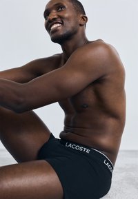 Black boxer briefs with a white "LACOSTE" logo on the waistband, shown on a male model with a muscular build against a neutral background.