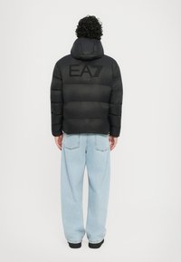 Schwarze Pufferjacke mit Kapuze und EA7-Logo auf dem Rücken, kombiniert mit hellblauen, oversized Jeans. Glatte Textur, gestepptes Design.