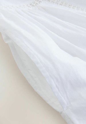 Material textil alb cu o dantelă texturată pe margine. Materialul pare ușor și ușor transparent, prezentând falduri moi și drapaje delicate.