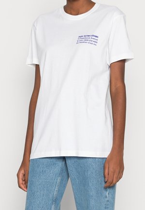 Valkoinen puuvillainen t-paita, jossa on lyhyet hihat, pyöreä pääntie ja vasemmassa rinnassa sininen painettu teksti, yhdistetty vaaleansinisiin farkkuihin.