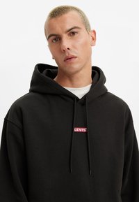 Sudadera con capucha negra hecha de una mezcla de algodón, que presenta un parche con el logo rojo de Levi's en el pecho y un cierre con cordón, con una textura suave.