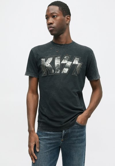 Camiseta gráfica negra con acentos desgastados, que presenta el logo de "KISS" en tachuelas plateadas. Cuello redondo y mangas cortas, combinada con jeans azules.