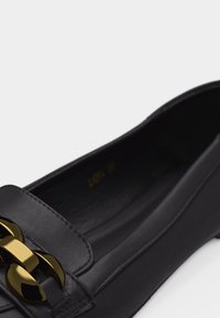 Mocassin plat en cuir noir avec un détail de chaîne dorée épaisse sur le devant et un design à bout arrondi.