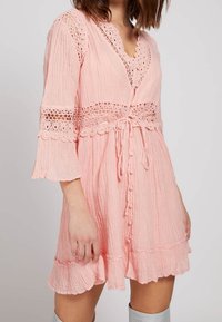Vestido rosa con textura, mangas de campana y acentos de encaje. Presenta un escote en V, cierre de botones y un detalle de cinturón en la cintura. Bajo con volantes.