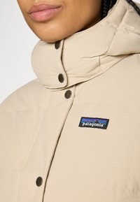 Detailný záber na osobu v béžovej puffer bunde Patagonia s čiernymi cvokmi a vysokým golierom, ktorý zakrýva krk.