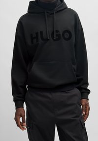 Sudadera negra hecha de tejido grueso con un bolsillo frontal. Presenta un gran logotipo "HUGO" mate en el pecho y cordones ajustables en la capucha.