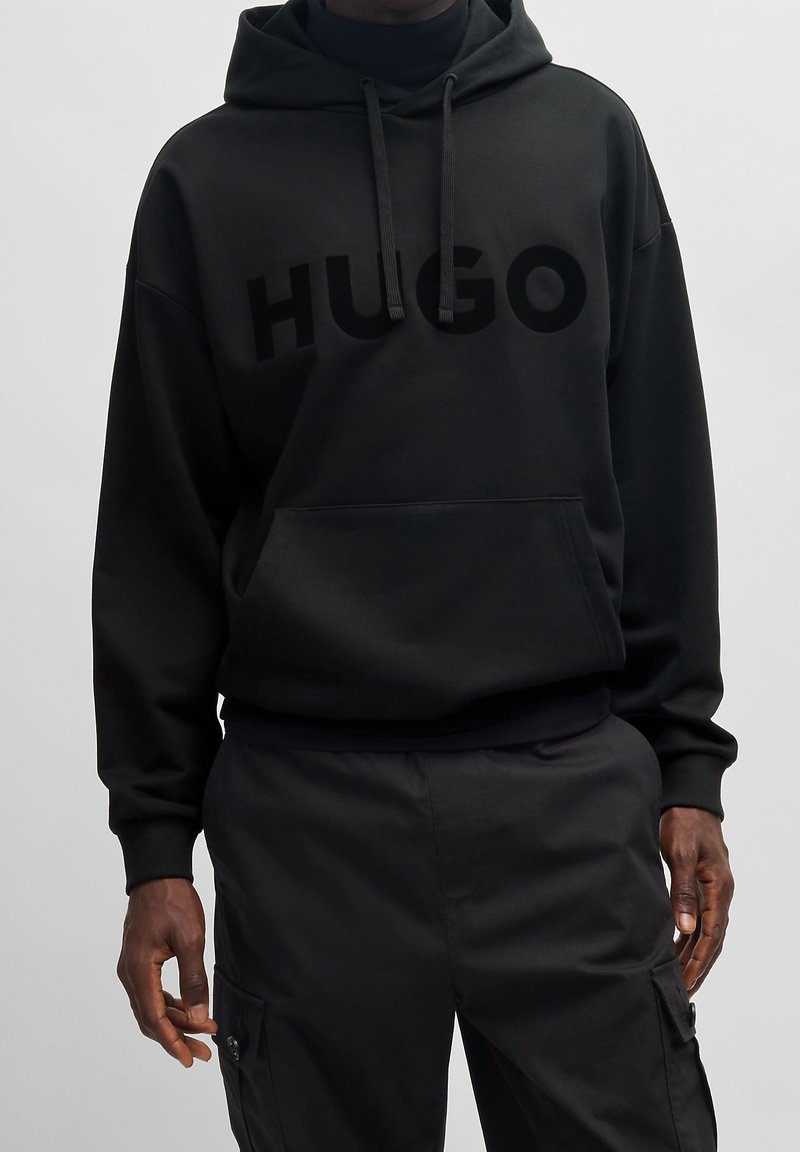 Sudadera negra hecha de tejido grueso con un bolsillo frontal. Presenta un gran logotipo "HUGO" mate en el pecho y cordones ajustables en la capucha.