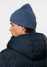 Blå strikket lue med ribbet tekstur og spiss topp, brukt sammen med en svart boblejakke med hette og quiltet design.
