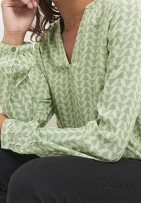 Lysegrøn bluse med geometrisk trekantmønster, med V-hals, rynkede ærmer og teksturdetaljer i stoffet.