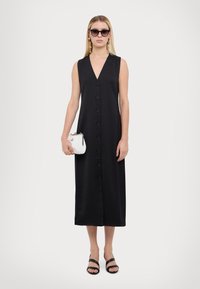Rochie neagră fără mâneci, cu un decolteu adânc în V, butoane în față și o croială lungă, vaporoasă. Asortată cu sandale negre și o geantă mică albă.
