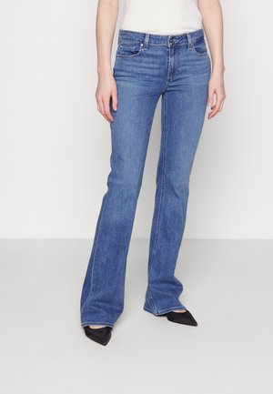 Personne portant un jean bootcut bleu taille moyenne et des chaussures noires à bout pointu, debout devant un fond clair uni.