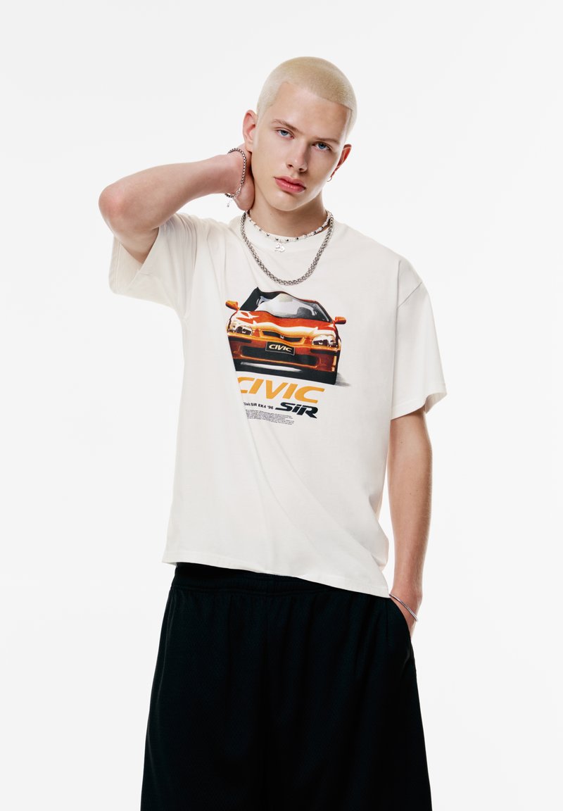 Creme t-shirt med grafisk print af en orange bil, med korte ærmer og afslappet pasform, parret med sorte shorts.