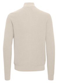 Pull en tricot beige avec un col montant et des manches longues. Texture côtelée, poignets et ourlet ajustés, et design dos sans couture. Pas de logos visibles.