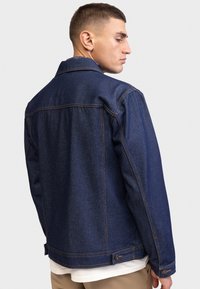 Chaqueta de mezclilla azul oscuro con un diseño clásico, con costuras naranjas, puños de botón y un corte relajado. La vista trasera muestra el cuello y las costuras.