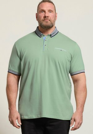 Mężczyzna z brodą w jasnozielonej koszulce polo z szarym kołnierzem i krawędziami rękawów, stoi na tle jednolitego beżowego tła.