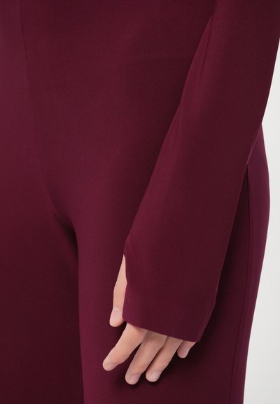 Bordové priliehavé oblečenie s dlhými rukávmi; má hladkú, elastickú látku a výrez na zápästí. Súčasťou je detailný záber ruky.