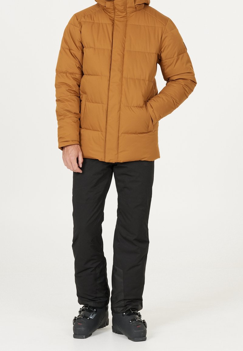 Homme portant une veste matelassée marron, un pantalon de neige noir et des chaussures de ski noires, debout avec une main dans la poche de la veste.