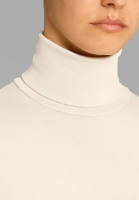 Maglione a collo alto color crema in un morbido tessuto a coste con cuciture curate e un colletto aderente. Primo piano della scollatura e dell'area della parte inferiore del viso.
