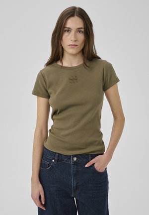 Nuori nainen, jolla on pitkät ruskeat hiukset ja päällään oliivinvihreä lyhythihainen paita sekä tummansiniset farkut, seisoo yksinkertaisen taustan edessä.