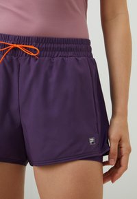 Fialové športové šortky s elastickým pásom a oranžovým šnúrkou. Majú malé logo na boku a hladký, ľahký materiál.