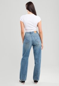 Jeans a vita alta di colore azzurro chiaro con gambe diritte, tasche posteriori e una toppa in pelle marrone in vita, abbinati a una t-shirt bianca aderente.