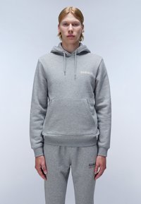 Sweat à capuche gris en tissu doux, présentant une poche kangourou, des cordons de serrage et un détail de logo en haut à gauche. Accompagné d'un pantalon de jogging gris.