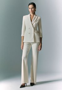 Blazer doppiopetto crema con maniche a tre quarti, abbinato a pantaloni a palazzo coordinati. I dettagli comprendono rever affilati e una vestibilità strutturata.