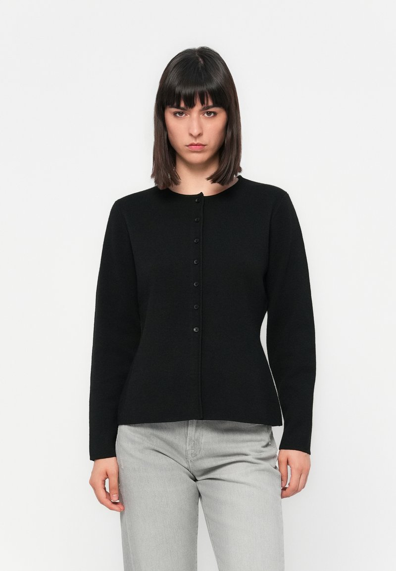 Kobieta z prostymi ciemnymi włosami, ubrana w czarny kardigan na guziki i jasnoszare dżinsy, stojąca na tle białej ściany.