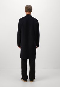 Man with short light brown hair wears long black coat, black pants, and black sneakers with white stripes, standing facing a white wall.