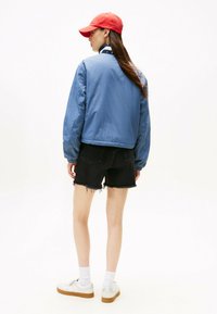 Jasnoniebieska kurtka bomberka o gładkim wykończeniu, czarne zniszczone szorty dżinsowe, białe sneakersy i czerwona czapka. Ogólnie swobodny styl.