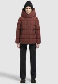 Braune gepolsterte Jacke mit einer breiten Kapuze, horizontaler Steppoptik, seitlichen Reißverschlüssen und schwarzen Akzenten. Kombiniert mit einer taillierten schwarzen Hose und Stiefeln.