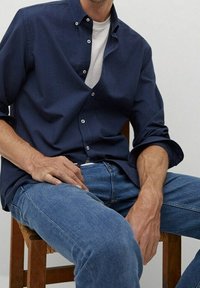 Granatowa koszula zapinana na guziki, wykonana z gładkiego materiału, noszona na białej koszulce. Połączona z niebieskimi dżinsami, siedzi na drewnianym krześle.