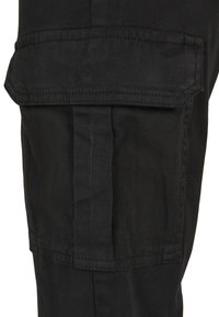 Černá látková nákladní kapsa s klopou a zesíleným prošitím, umístěná na kalhotách.