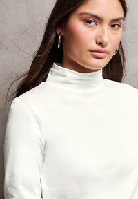 Witte lange mouw top met hoge hals en gladde textuur; model draagt zilveren creolen; neutrale achtergrond.