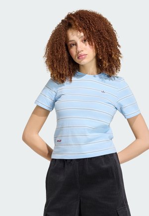 Joven con cabello rizado rojo, que lleva una camisa de manga corta a rayas azul claro y pantalones de pana negros, de pie con las manos detrás de la espalda.