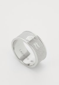 Bague en argent avec une surface texturée présentant un motif subtil et un logo. Anneau intérieur lisse avec le nom de la marque affiché à l'intérieur.