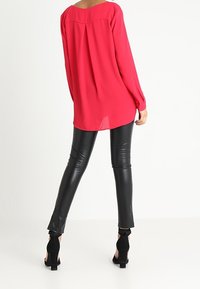 Blusa rossa in chiffon con una vestibilità morbida, maniche lunghe e piega sul retro, abbinata a pantaloni in pelle nera attillati e scarpe con tacco alto nere.